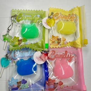 May include: Four colourful candy-shaped toys in clear plastic pouches. Each pouch has a zip closure and a keychain. The candies are green, yellow, blue, and pink. Each pouch has the word "Candy" printed on it.