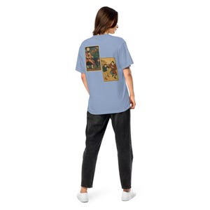 以下が含まれることがあります： 背中に2つの長方形のイラストが描かれた水色のTシャツ。イラストは伝統的な衣装を着た人物を描いています。その人は黒いジーンズと白いスニーカーを履いています。