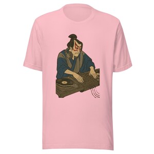 以下が含まれることがあります： 日本の侍DJのグラフィックがプリントされたライトピンクのTシャツ。侍はヘッドホンを着用し、ターンテーブルを操作しています。グラフィックは茶色、青、赤の色合いです。
