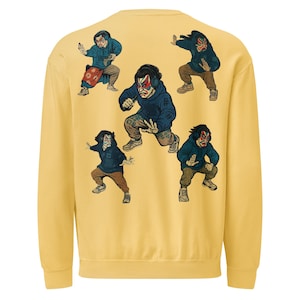 以下が含まれることがあります： マスタードイエローのスウェットシャツ。6人の人物がアクションポーズをとるグラフィックが特徴です。各人物はマスクと青いパーカーを着用しています。人物は様々な姿勢で、詳細な表情と服装をしています。