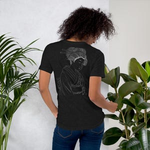 以下が含まれることがあります： 背中に白い線画の芸者の絵が描かれた濃いグレーのTシャツ。 芸者は伝統的な着物と髪型をしています。 シャツは柔らかい素材でできており、クラシックなクルーネックです。
