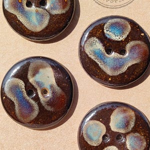 Könnte beinhalten: Vier runde Knöpfe mit zwei Löchern und einer glänzenden dunkelbraunen Glasur mit irisierenden blauen und cremefarbenen abstrakten Mustern. Ein kreisförmiger Stempel auf der Rückseite zeigt Handmade ELIZA'S BUTTONS in Oxford. Die Knöpfe sind glatt.