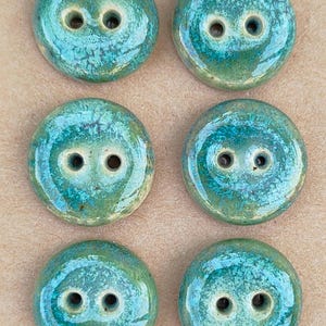 May include: Six handmade, round ceramic buttons in teal and green hues. Each button features two sewing holes. The buttons are displayed on a tan card, with a circular stamp reading "Handmade ELIZA'S BUTTONS in Oxford".