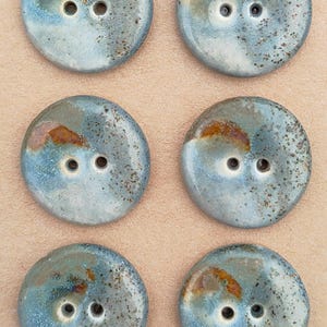 Könnte beinhalten: Ein Set aus sechs runden Keramikknöpfen mit jeweils zwei Löchern, die eine glänzende Glasur in Blau-, Grau- und Brauntönen mit gesprenkelten Details aufweisen. Die Knöpfe sind in zwei Spalten auf einer hellbraunen Karte mit dem Wort Handmade angeordnet.