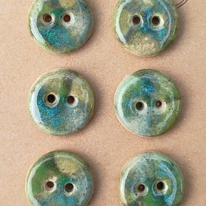 Peut inclure: Six boutons ronds faits à la main, chacun avec deux trous, disposés sur une carte beige. Les boutons présentent une finition brillante avec un mélange de couleurs vertes, bleues et dorées. La carte est estampillée "Handmade Eliza's Buttons".