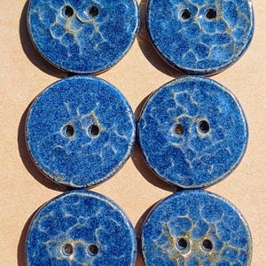 May include: A set of six round, two-hole buttons featuring a deep blue glaze with a textured, crackled finish. The buttons are arranged in two vertical columns of three on a light brown card, displaying a glossy surface with subtle variations.