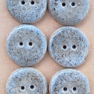 Puede incluir: Seis botones redondos de dos agujeros con un acabado brillante gris claro moteado. Los botones están dispuestos en dos columnas de tres sobre una tarjeta de color canela con un sello circular que dice Handmade ELIZA'S BUTTONS en tinta negra.