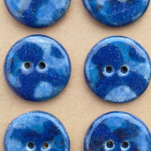 May include: A set of six round, glossy ceramic buttons arranged in two columns on a tan card. Each button features a mottled blue and dark navy glaze with two central holes. The top right corner of the card displays a circular stamp reading handmade.
