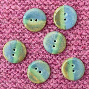 May include: Six round, two-hole buttons featuring a smooth, glazed finish with a color gradient transitioning from blue to green and earthy brown tones. Each button displays a unique blend of these colors, arranged in a circular pattern.