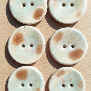 May include: A set of six round ceramic buttons arranged in two columns on a rectangular cardboard backing. Each button features a speckled light blue and cream glaze with irregular brown spots and two central holes. The text Handmade ELIZA'S is visible.