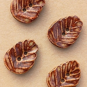 May include: Four brown ceramic buttons shaped like leaves with a glossy finish and two holes in the center. The buttons are attached to a tan card featuring a circular stamp that reads Handmade ELIZA'S BUTTONS in Oxford.