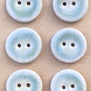 Könnte beinhalten: Sechs runde Keramikknöpfe in zwei Spalten, jeder mit zwei Löchern und einem Farbverlauf aus hellblauer und weißer Glasur. Die Knöpfe sind auf einer beigen Karte mit dem in schwarzer Tinte gestempelten Text Handmade ELIZA'S BUTTONS befestigt.