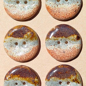 May include: A set of six round ceramic buttons arranged in two columns on a tan card with the word Handmade stamped in the corner. Each button features a glossy finish with horizontal bands of dark brown, muted blue-gray, and speckled orange.