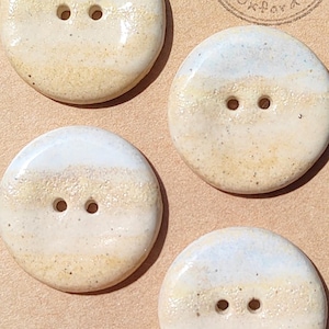 May include: Four round, two-hole ceramic buttons with a glossy finish, featuring horizontal bands of cream, beige, and light blue speckles. The buttons are attached to a tan card stamped with the text Handmade ELIZA'S BUTTONS in Oxford.