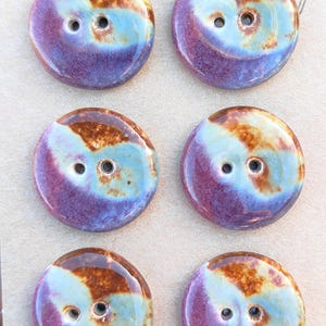 May include: A set of six round ceramic buttons arranged in two columns on a light tan card. Each button features a glossy, multi-colored glaze with swirling patterns of deep purple, light blue, and warm brown. Each button has two central holes.