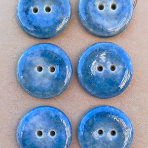Könnte beinhalten: Ein Set aus sechs runden Zwei-Loch-Knöpfen mit einer glänzenden, gesprenkelten blauen Glasur und variierenden Farbintensitäten. Die Knöpfe sind in zwei Spalten zu je drei Stück auf einer hellbraunen Karte angeordnet, auf der das Wort Handmade steht.