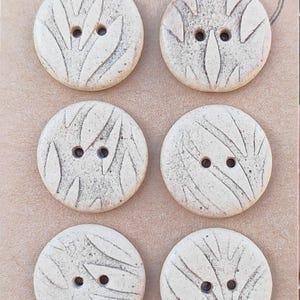 May include: A set of six round, off-white buttons arranged in two columns on a tan card. Each button features two centre holes and a textured, carved leaf-like pattern with dark accents. A circular stamp in the top right corner reads Handmade ELIZA'S.