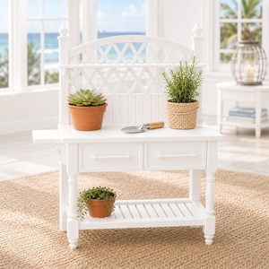 May include: A white potting bench with a lattice back, two drawers, and a lower shelf. It holds potted plants, a trowel, and a woven basket. The bench is set on a woven rug, with a beach scene visible in the background.