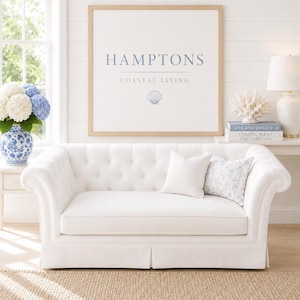 May include: A white tufted sofa with two pillows, one solid white and one patterned. A framed print above the sofa reads "HAMPTONS COASTAL LIVING". A vase of blue and white flowers sits to the left.
