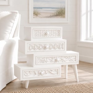 Puede incluir: Mesa auxiliar blanca con cuatro cajones, cada uno con diseños tallados únicos. Los cajones incluyen patrones tejidos, circulares y florales. La mesa tiene cuatro patas cónicas y está colocada sobre una alfombra tejida. La mesa mide 76 cm de alto.