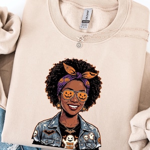 May include: A sand-colored crewneck sweatshirt featuring a graphic illustration of a person with curly hair wearing a purple headband with pumpkin patterns, aviator sunglasses with pumpkin reflections, and a denim jacket with ghost and bat motifs.