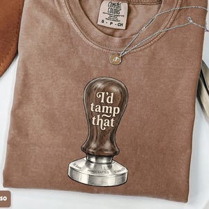 May include: Espresso-colored t-shirt with the phrase "I'd tamp that" printed on the front, featuring a coffee tamper graphic. The shirt has a crew neck and short sleeves. A silver necklace with a pendant is draped across the shirt.