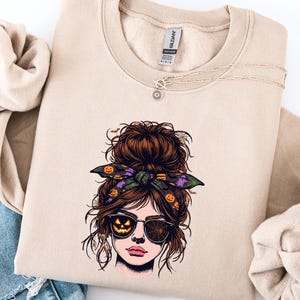 May include: A sand-colored crewneck sweatshirt featuring a graphic print of a person with a messy bun hairstyle, wearing a headband with pumpkin patterns and sunglasses reflecting a jack-o'-lantern and a spider web design.