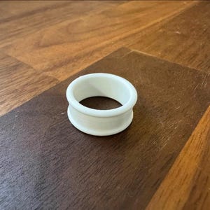 以下が含まれることがあります： 中央に穴のある、白く円筒形の物体。物体は滑らかでマットな表面を持っています。木製の表面に置かれています。