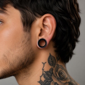 May include: A solid black circular ear tunnel plug inserted into a stretched earlobe. The jewelry features a smooth, matte finish with a hollow center, designed to sit flush against the skin of the earlobe, providing a clean and simple aesthetic.