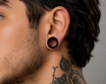 Puede incluir: Un túnel de oreja circular negro sólido con un acabado liso y mate. La joya presenta un centro hueco y un borde frontal ancho y plano, diseñado para insertarse en una perforación de lóbulo estirada para una apariencia limpia y uniforme.