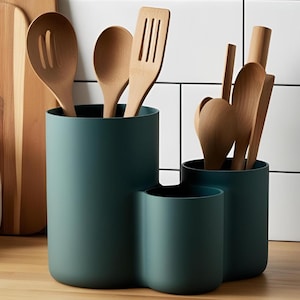 Puede incluir: Soporte para utensilios de cocina de color verde azulado con tres compartimentos, lleno de cucharas de madera, espátulas y otros utensilios de cocina. El soporte está sobre una superficie de madera, con una tabla de cortar y un tarro en el fondo.