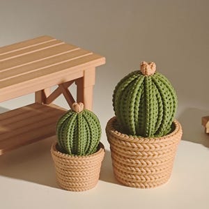 Puede incluir: Dos esculturas de cactus verdes en macetas de estilo tejido. Los cactus tienen un aspecto texturizado, con un pequeño detalle marrón claro en la parte superior. Un pequeño banco de madera y una paleta de madera en miniatura están en el fondo.