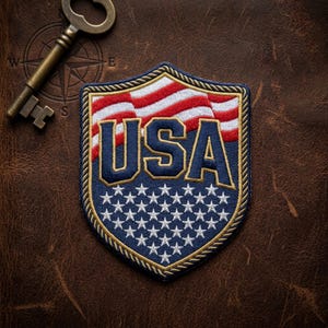 May include: Embroidered shield-shaped patch featuring the word "USA" in navy blue, with a waving American flag design in red and white. The patch has a border of gold stitching and a field of white stars on a blue background.