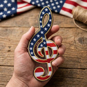 May include: A patriotic music-themed patch in the shape of a treble clef. The top half features a blue background with white stars, while the bottom half displays red and white stripes. The patch is outlined in gold and has a white border.