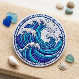 May include: Embroidered patch featuring a stylized ocean wave design in shades of blue and white. The circular patch has a white border with a rope-like texture. The wave design includes intricate details and a sense of movement.