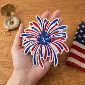 May include: A patriotic embroidered patch in the shape of a firework, featuring red, white, and blue colors. The patch has a starburst design with star and sequin accents. A small American flag is visible in the background.