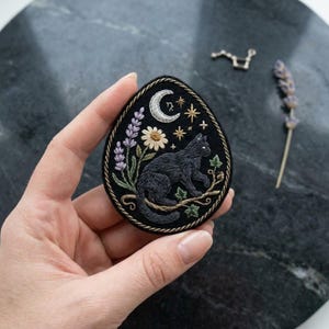 May include: An egg-shaped black fabric patch featuring detailed embroidery of a black cat perched on a branch, surrounded by lavender sprigs, a white daisy, a crescent moon, and small gold stars, all bordered by a twisted gold metallic cord trim.