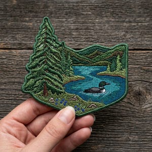 May include: An embroidered patch featuring a detailed landscape scene with a tall evergreen tree on the left, a blue lake in the center, and a loon swimming on the water. The background shows rolling green hills and mountains under a clear sky.