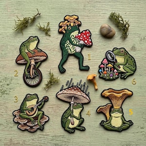 May include: Embroidered patches featuring whimsical frog designs. One frog rides a unicycle on a mushroom, another plays a lute, and others hold mushrooms or use them as umbrellas. The patches are green, brown, and red.