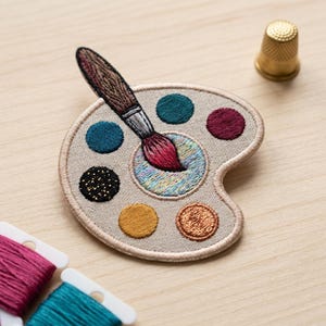 May include: An embroidered patch shaped like an artist's palette in beige, featuring a paintbrush with a red tip resting on a central mixing area. The palette includes circular paint spots in teal, burgundy, black with gold flecks, mustard, and copper.