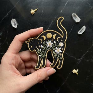May include: An embroidered cat-shaped patch featuring a black base with gold-colored moon phases across the back, white floral embroidery with green stems, and small gold stars. The patch has a gold-colored border and a curved, upright tail.
