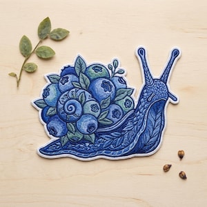 May include: Embroidered patch featuring a blue snail with a shell made of blueberries and green leaves. The snail has detailed stitching and a white border. A sprig of leaves and a few seeds are also visible.