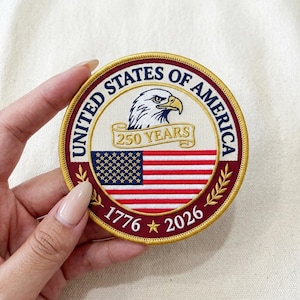 May include: A round embroidered patch with a patriotic theme. The patch features the text "UNITED STATES OF AMERICA" around the edge, an eagle, a banner reading "250 YEARS", and the American flag. The years "1776" and "2026" are also displayed.
