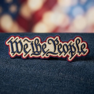 May include: Embroidered patch with the words "We the People" in a stylized font. The patch features a blue background with red and cream-colored accents. The patch is on a blue denim surface.