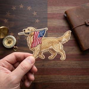 May include: Embroidered patch featuring a golden retriever dog carrying a small American flag in its mouth. The flag has red and white stripes and a blue field with white stars. The patch is held in front of a wooden surface.