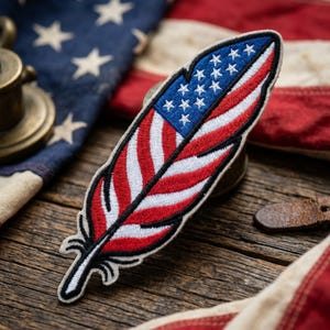 May include: Embroidered patch in the shape of a feather, featuring the American flag design. The patch has a blue section with white stars and red and white stripes. The patch is on a wooden surface with a vintage American flag in the background.