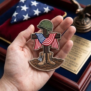 May include: Embroidered patch featuring a cross with a helmet, American flag, and combat boots. The patch is brown, green, red, white, and blue. The patch is held in a hand. A folded American flag and a bronze eagle statue are in the background.