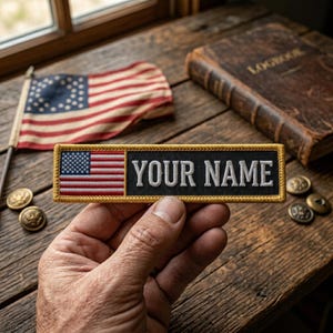 May include: A rectangular patch with an American flag design and the text "YOUR NAME" in white lettering. The patch is held in a hand, with a vintage American flag, buttons, and a book in the background. The patch has a black background and a gold border.