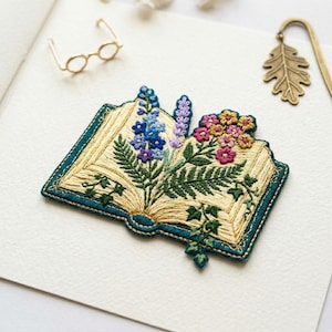 May include: Embroidered open book with colorful flowers and foliage. The book has a teal border and is filled with detailed stitching in various colors. A gold-colored bookmark and small glasses are also visible.