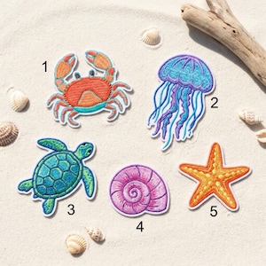 May include: Embroidered patches featuring sea creatures: a crab, jellyfish, sea turtle, seashell, and starfish. The patches are in vibrant colors, including orange, blue, green, and purple. The patches are on a sandy background with seashells and driftwood.